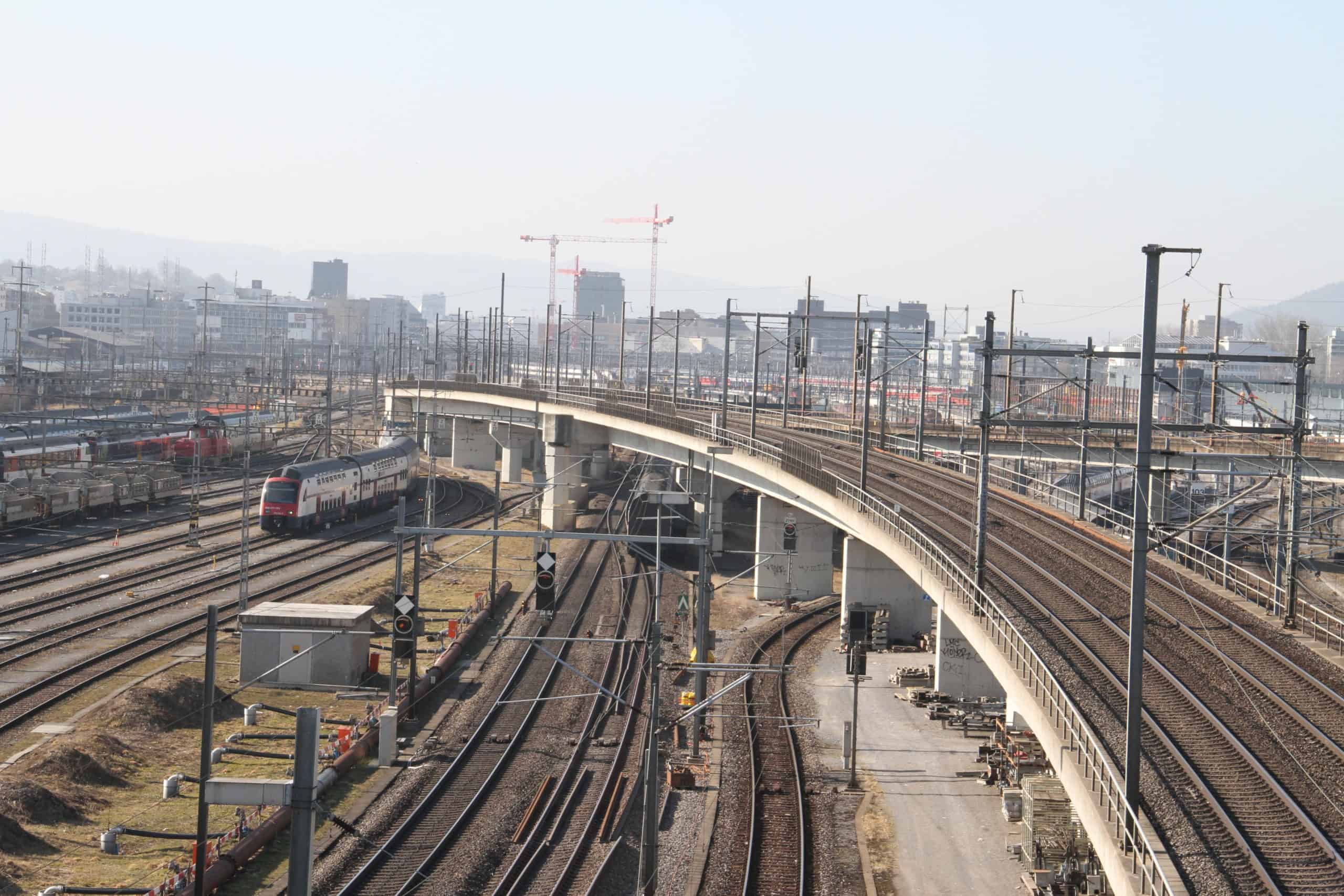 several high-speed trains on tracks at a busy junction and railway bridge in the foreground and a large city with high-rise buildings and cranes in the background