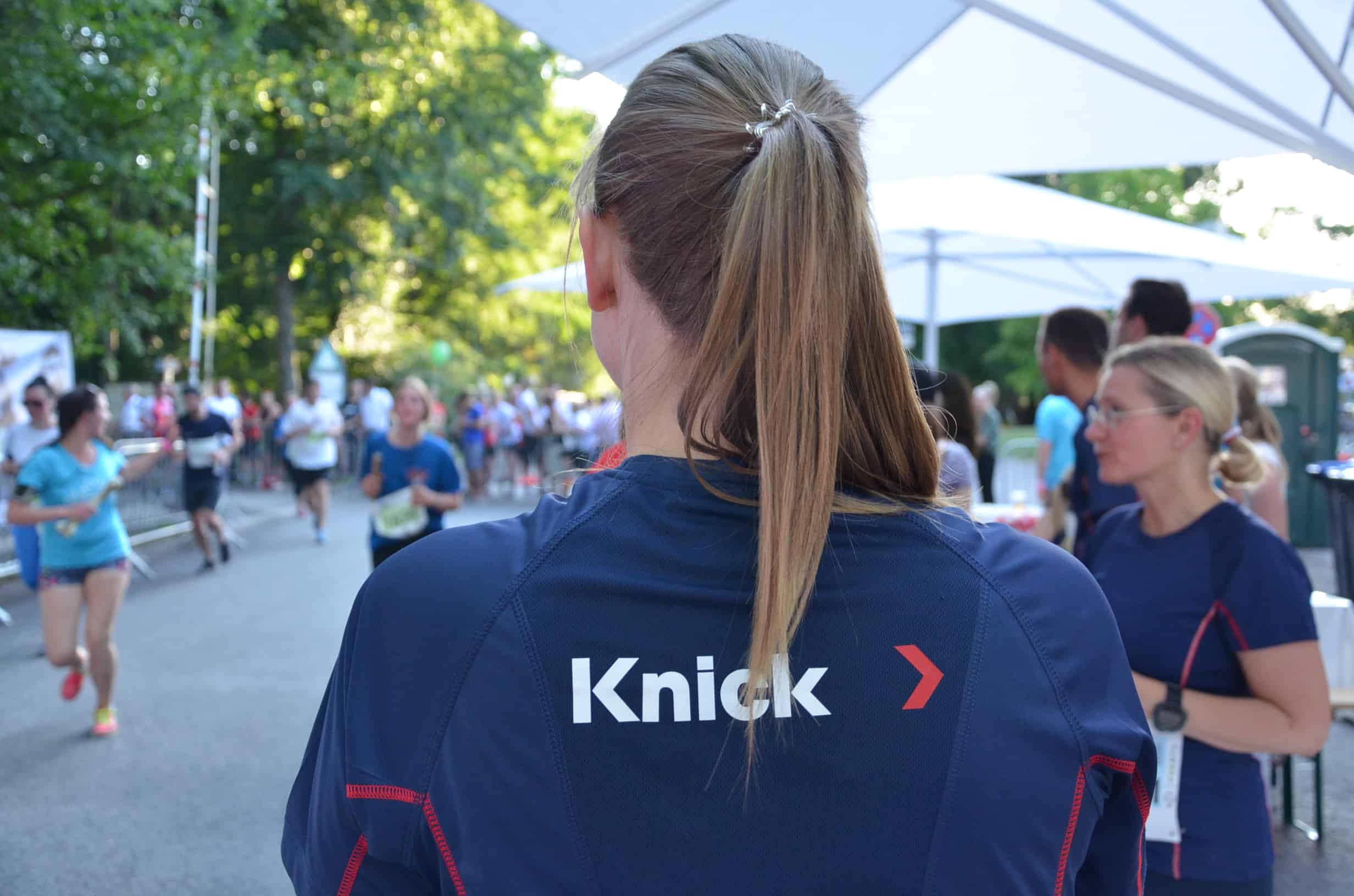 A female employee with ponytail and blue runner's shirt with the Knick logo on the back watches runners at a relay race in Berlin under large white umbrellas