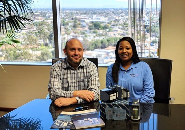 Two smiling Knick interface employees, a man in a checked shirt and a young woman in a blue blouse sitting at a table with electrical measuring devices and product brochures in front of them. they are sitting a bright office with a large window showing a skyline and a large palm tree outside